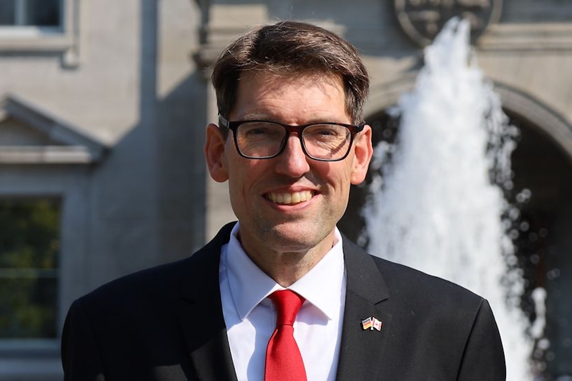 A head-and-shoulders shot of Matthias Lüttenberg. He is wearing a dark suit with a red tie and a lapel pin of the German and Canadian flags. There is a fountain in the background.