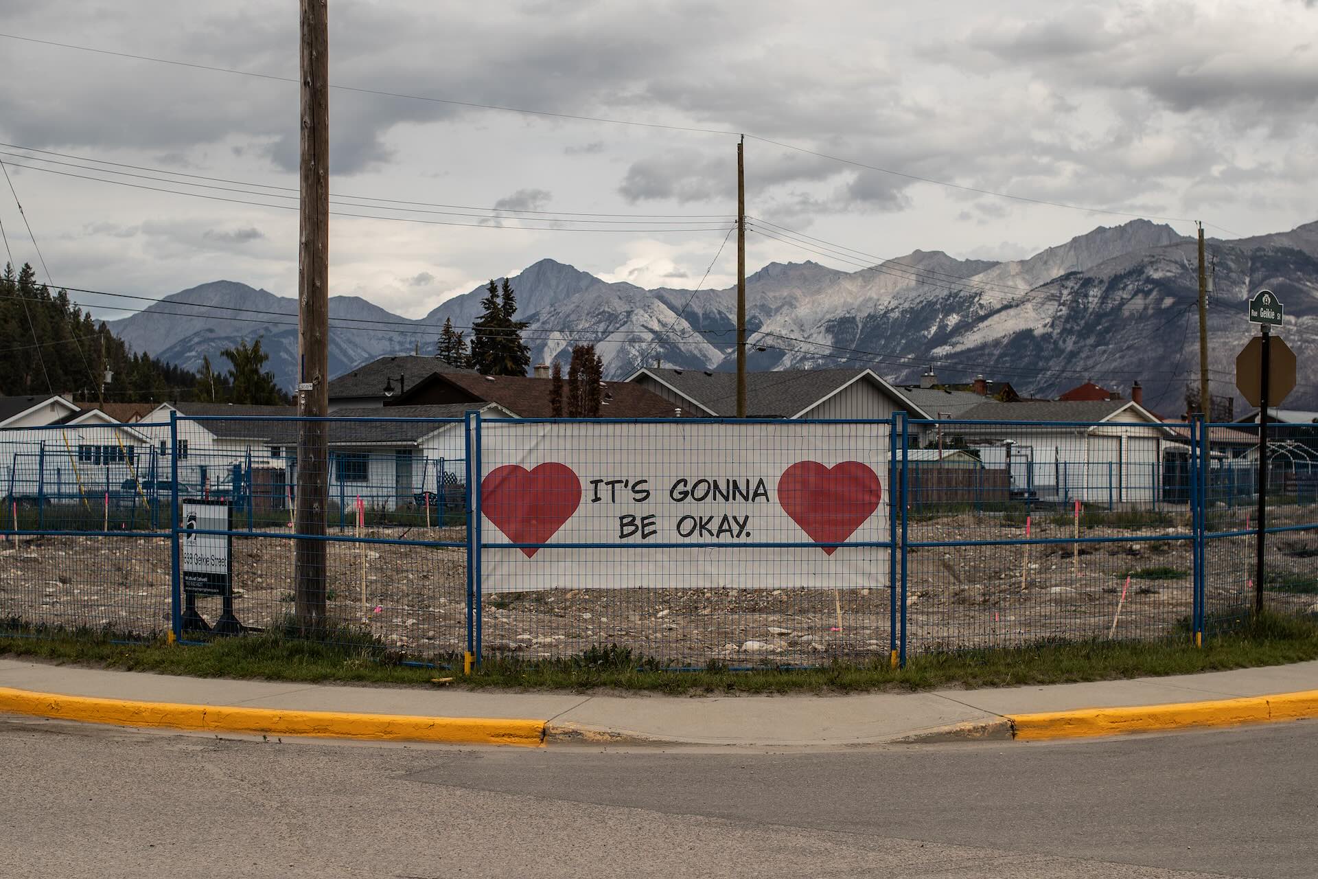 A sign reading "It's gonna be okay" with hearts on either side hangs from a blue wire construction fence around an empty lot at the corner of an intersection. Homes and mountains can be seen behind the lot.