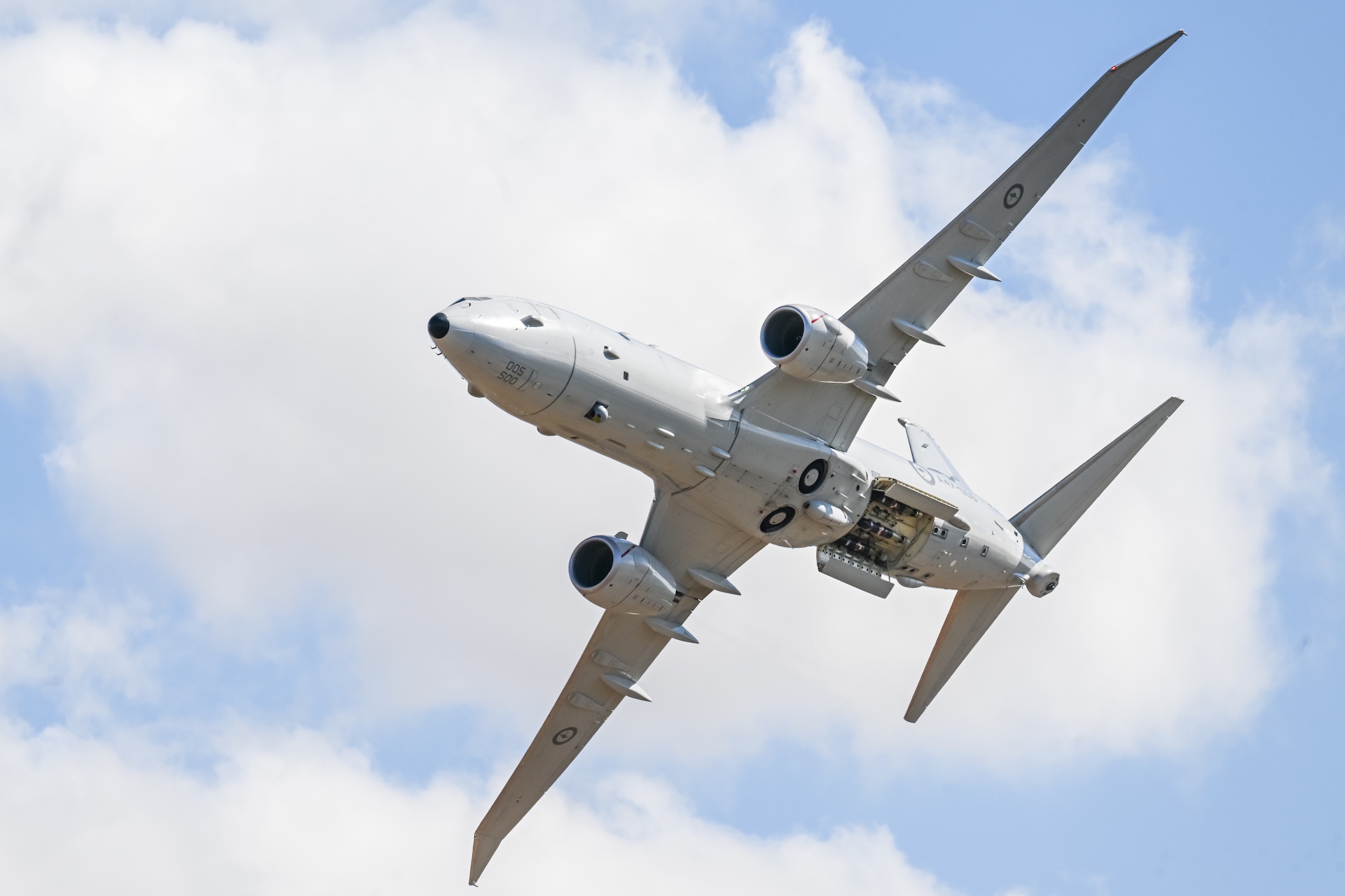 A shot from the ground of a large, twin engine jet flying overhead, with accordian-style doors on its undercarriage open.