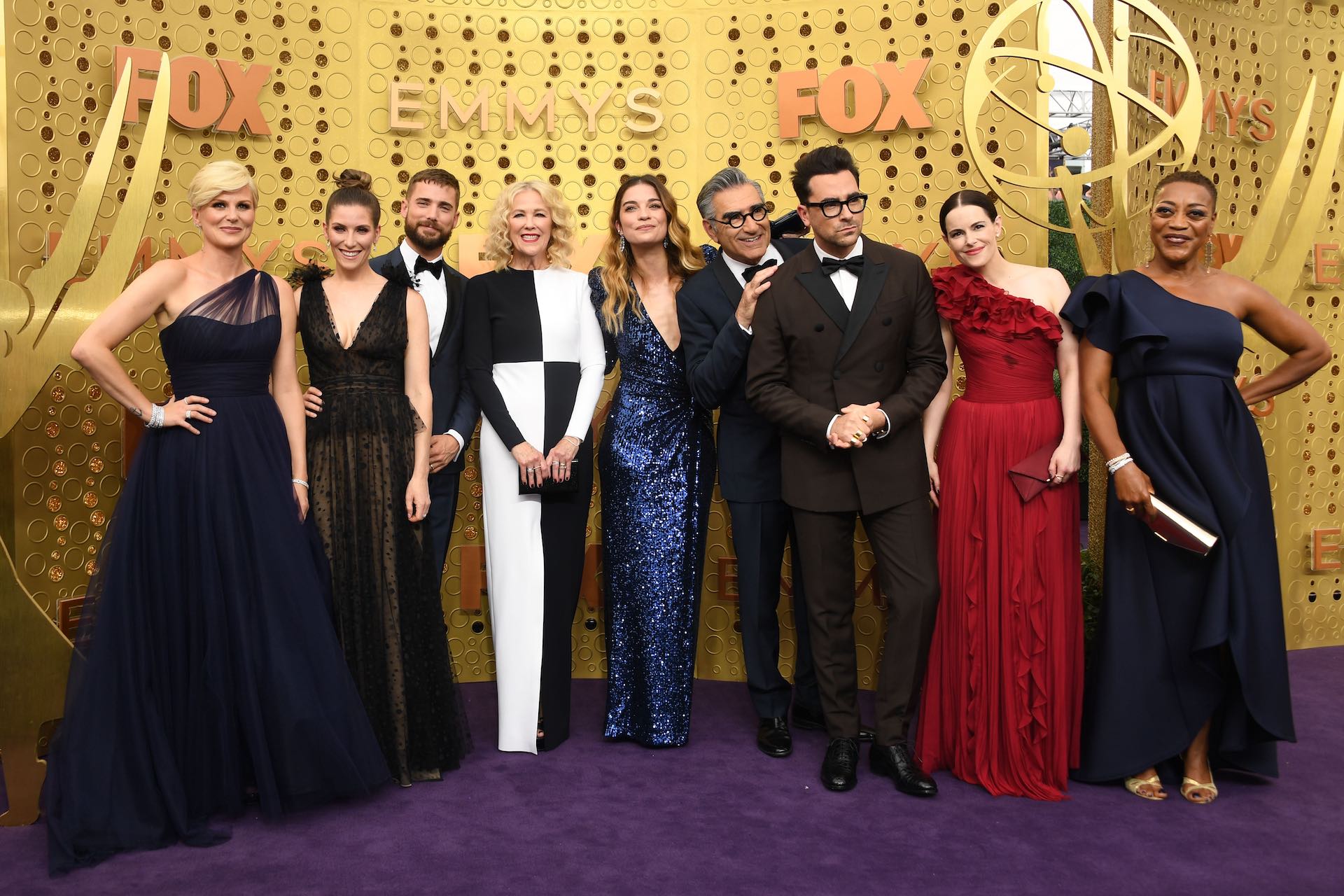 A party-style ensemble shot of nine cast members of Schitt's Creek in formal wear. They're standing in front of a gold backdrop with the word "Emmys" on it, and large figurines that resemble Emmy Awards trophies.