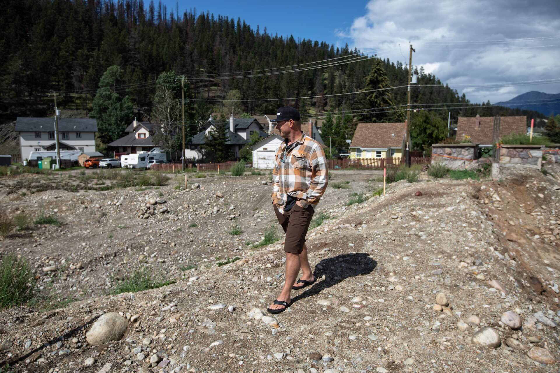 A man in sandals, shorts, a checkered shirt and a baseball hat walks with his hands in his pockets on a rocky lot of land. In the background across the street are a number of homes and trees.
