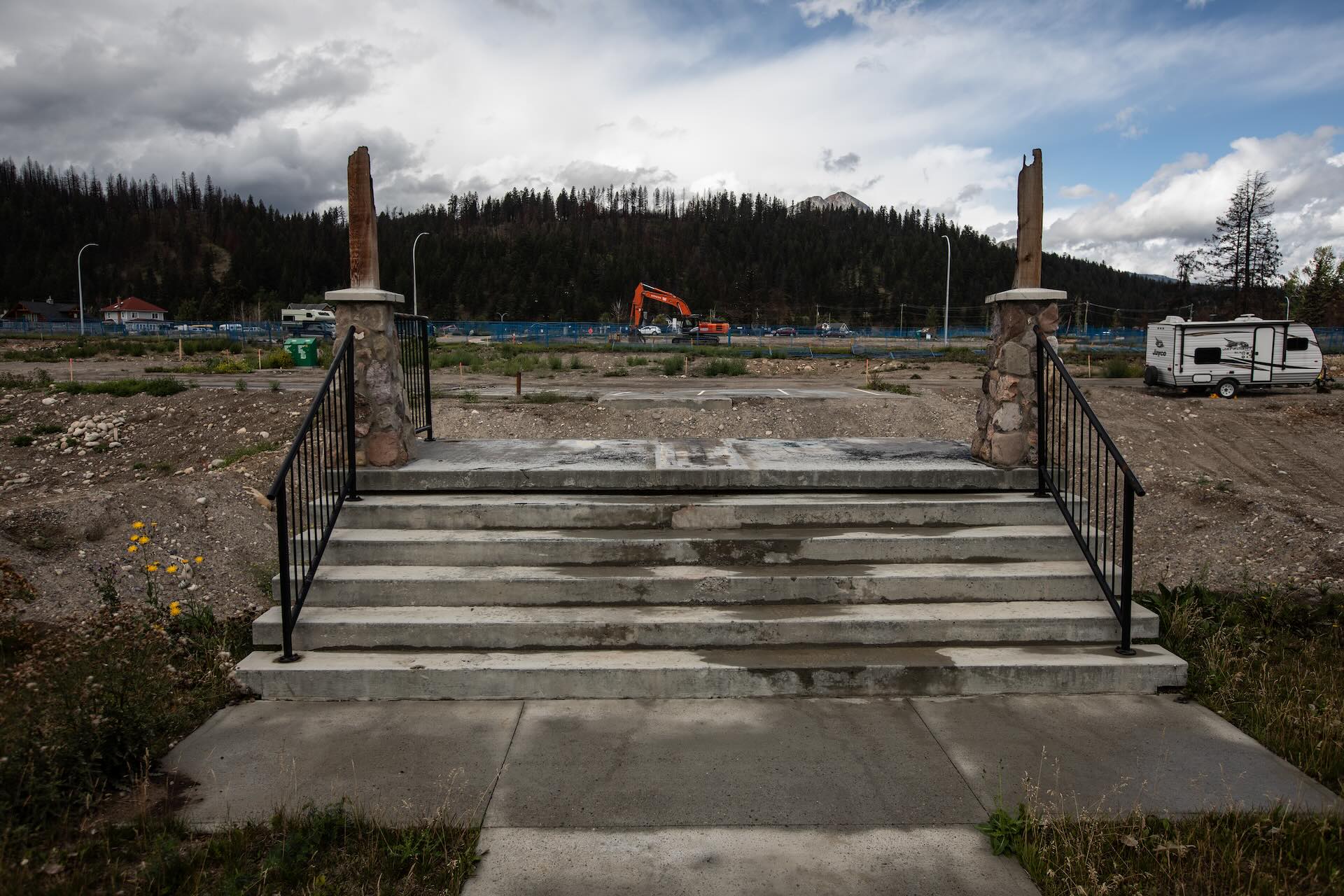 A set of steps lead up to stone pillars with broken wooden pillars on top of them. Behind the pillars is a vacant lot with some trucks and a crane around it.