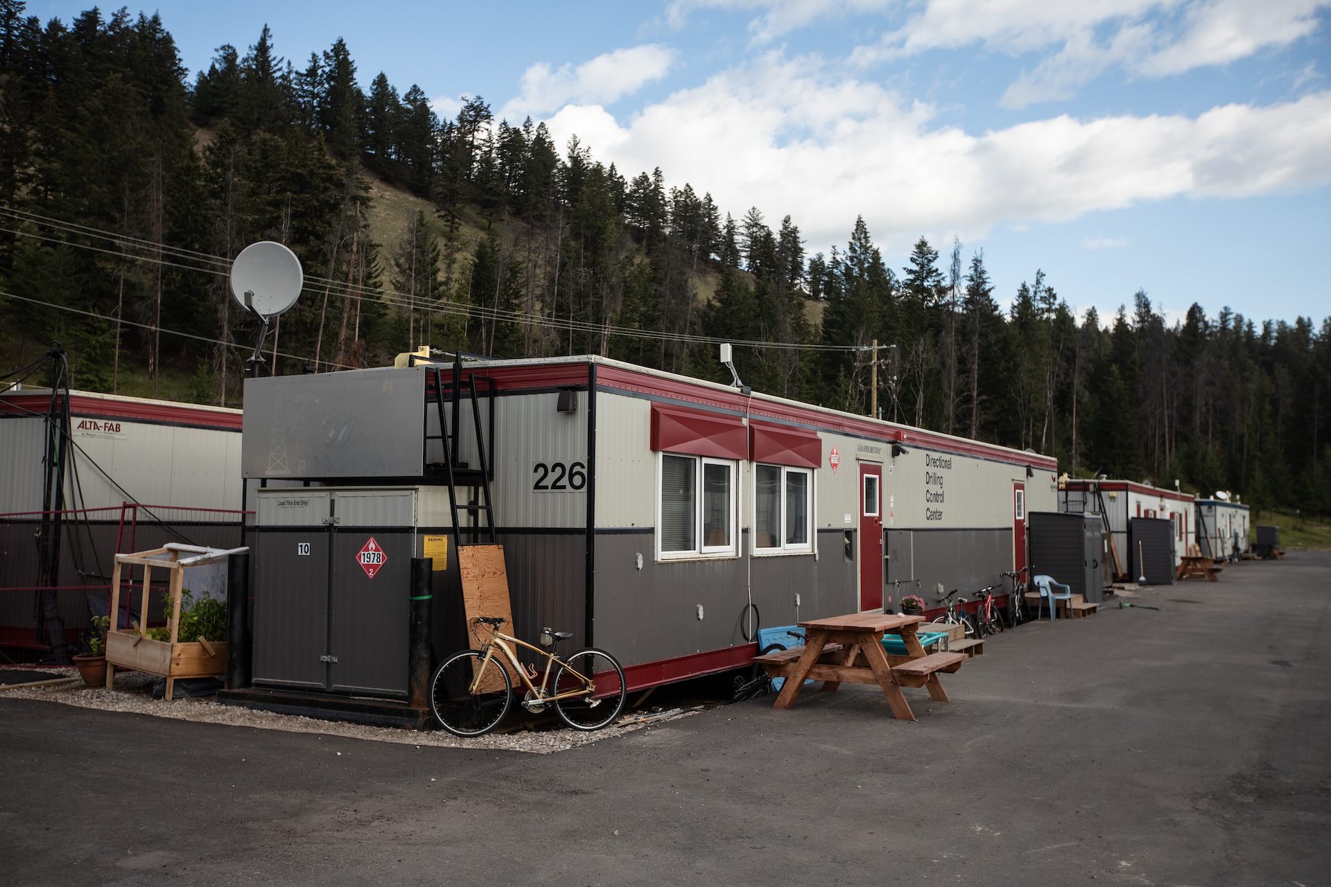 White-and-grey trailers with red doors and trim sit on an asphalt lot. There are park benches, bicycles, and a small standing herb garden around them, with a satellite dish on top of one trailer.