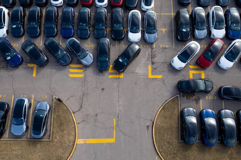 An aerial shot of a lot full of Tesla cars. The company's logo is painted in yellow on the asphalt.