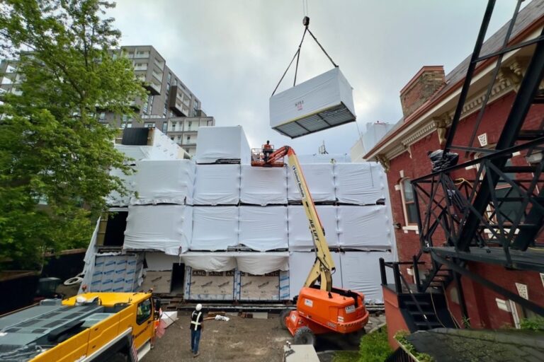Construction site with a crane lifting a large, wrapped module onto a stacked structure. Workers with safety gear, buildings, and trees in the background. Urban development scene.