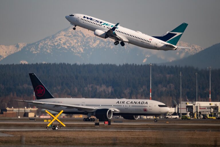 An Air Canada jet stands on the runway at an airport while a WestJet plane takes off above it and in the background