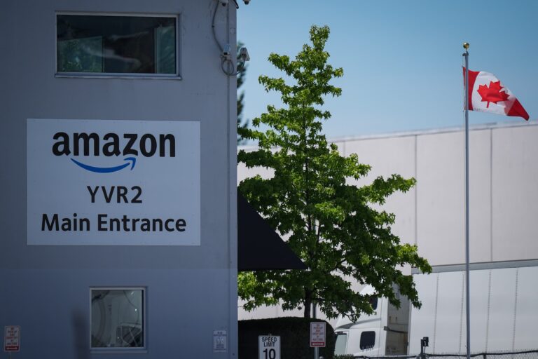 A building on the left of the frame has a sign reading "Amazon YVR2 Main Entrance." To the right of the building are a tree, a Canadian flag on a flagpole, and a transport truck.