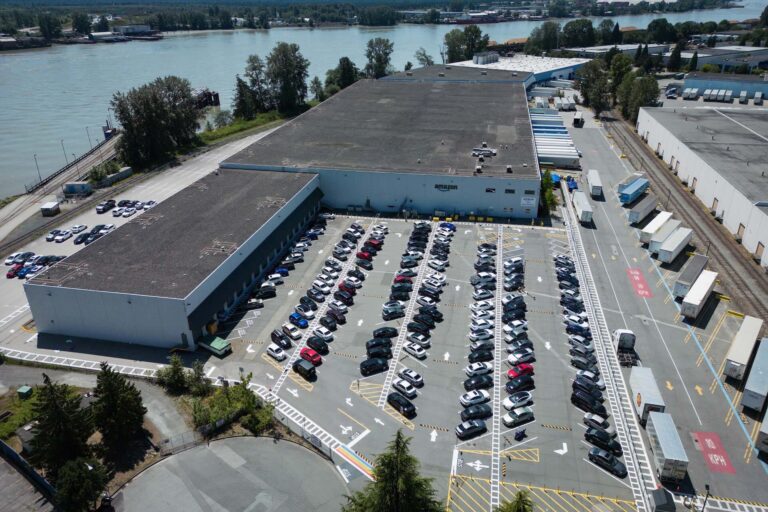 Aerial view of an Amazon warehouse and parking lot with a river alongside a river in the background.
