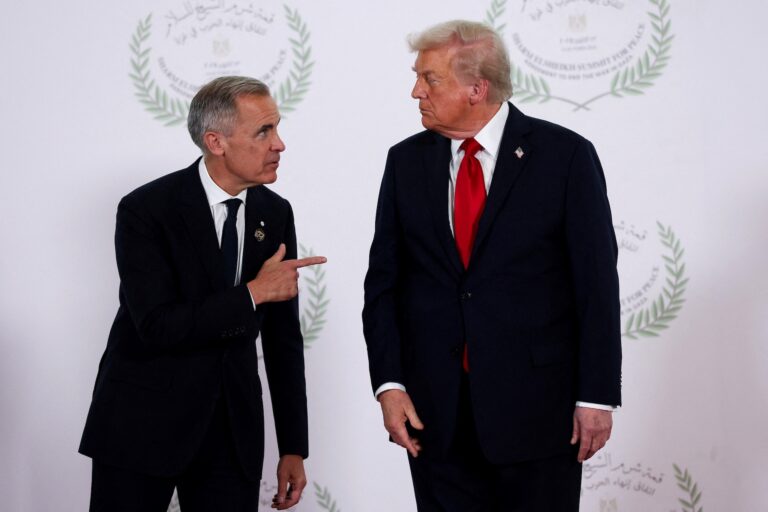 Carney and Trump at a photo op in Sharm El-Sheikh, Egypt, against a white backdrop that features a peace-themed logo for the gathering. Carney is leaning toward a scowling Trump and pointing his index finger at the U.S. president.