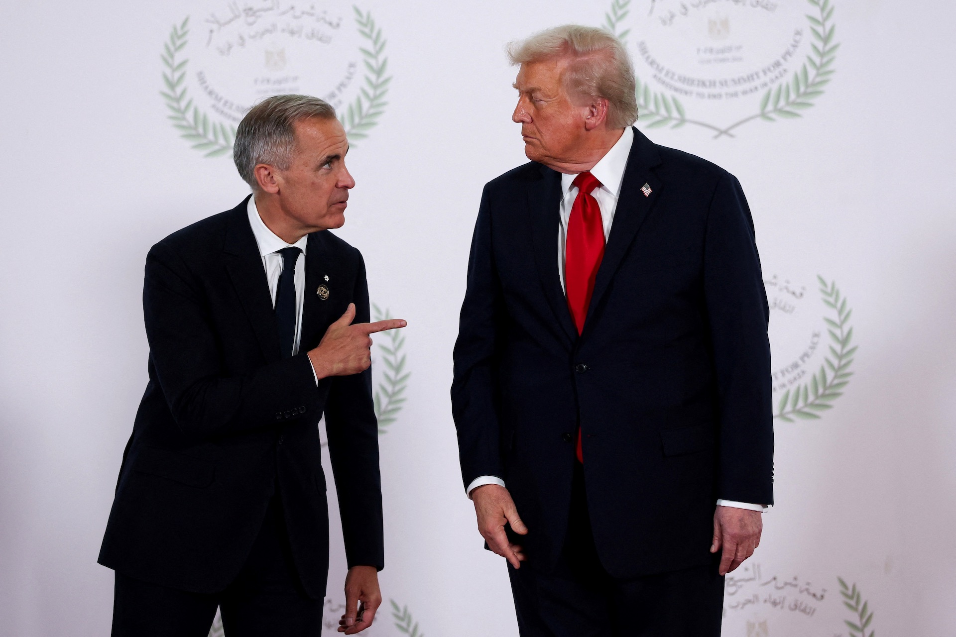 Carney and Trump at a photo op in Sharm El-Sheikh, Egypt, against a white backdrop that features a peace-themed logo for the gathering. Carney is leaning toward a scowling Trump and pointing his index finger at the U.S. president.