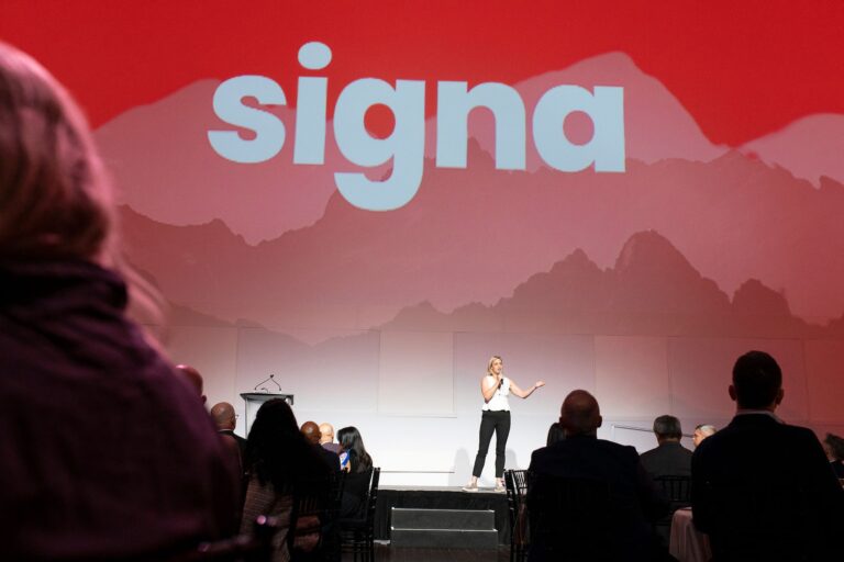 A shot down the centre aisle of an audience watching Dana O'Born on stage. O'Born is speaking into a microphone, and Signa's logo looms behind her on a towering red backdrop.