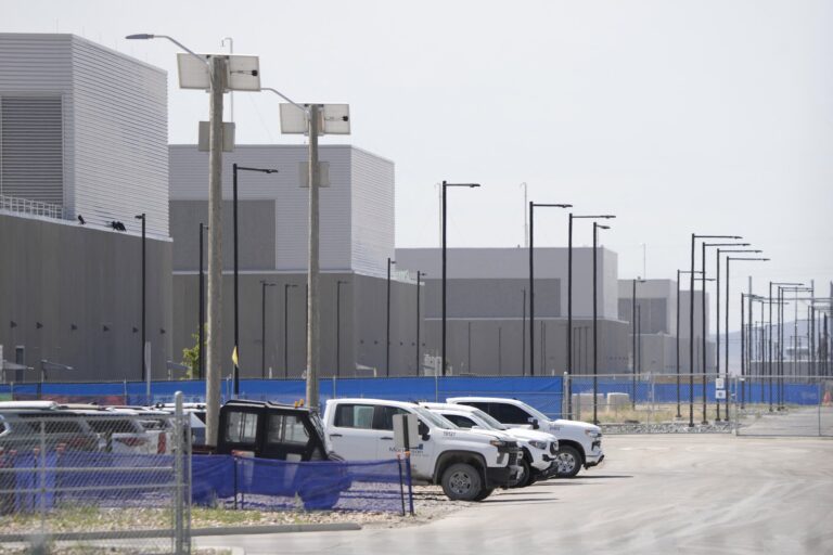 A wide shot of a rows of towering industrial-style buildings on the Utah desert. There are white pickup trucks parked in the foreground.