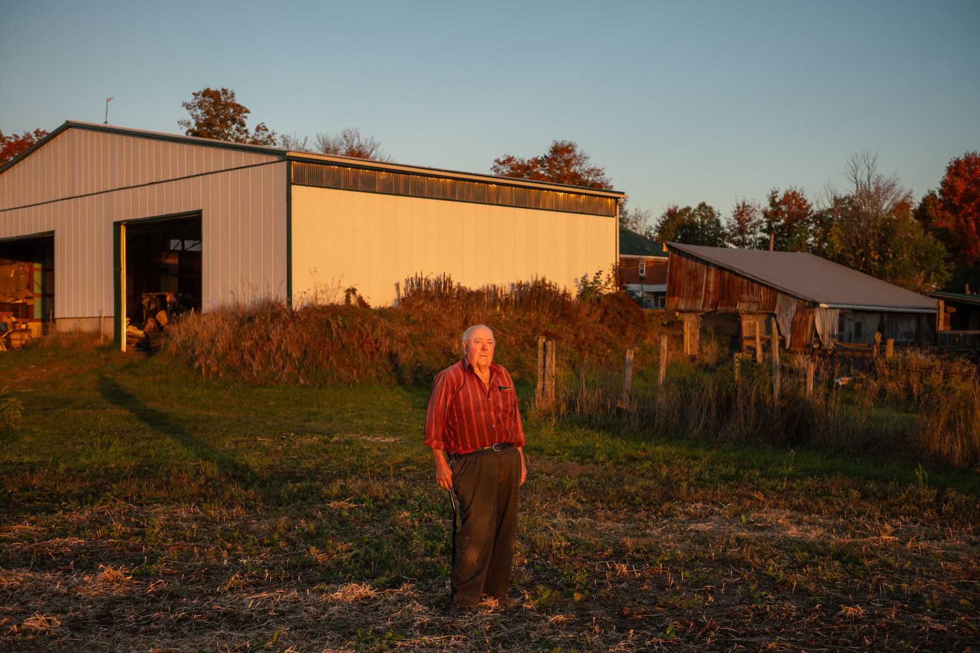 An older man in a red striped shirt and dark slacks stands in a field looking at the camera, with farm buildings in the background.