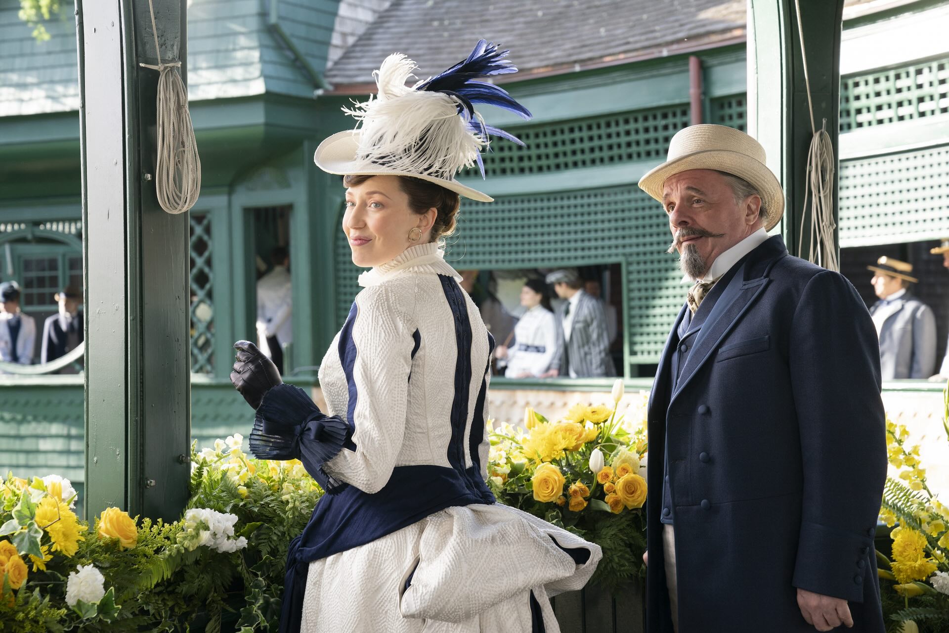 A woman in a fancy white and blue dress with an ornate matching hat stands next to a many in a blue suit and hat in a late-1800s-set scene from the HBO series The Gilded Age.