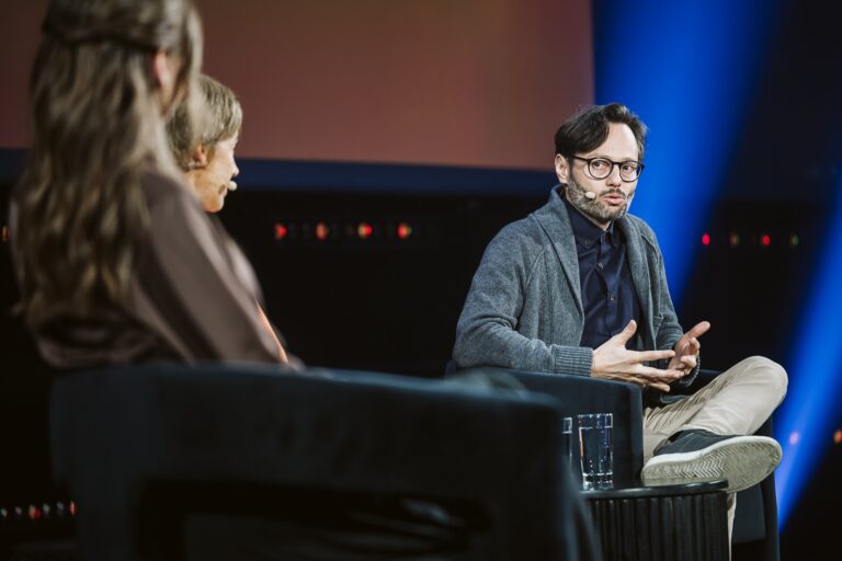 A man in a blue shirt and grey sweater sits in a chair on stage and speaks to two other people seated off to his right.