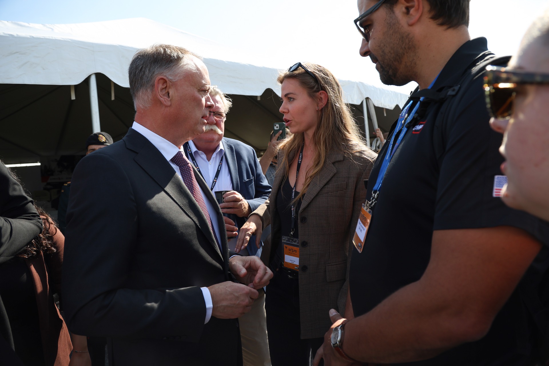 A shot of Katheron Intson speaking with David McGuinty amid a group of people. There is a white awning in the background.