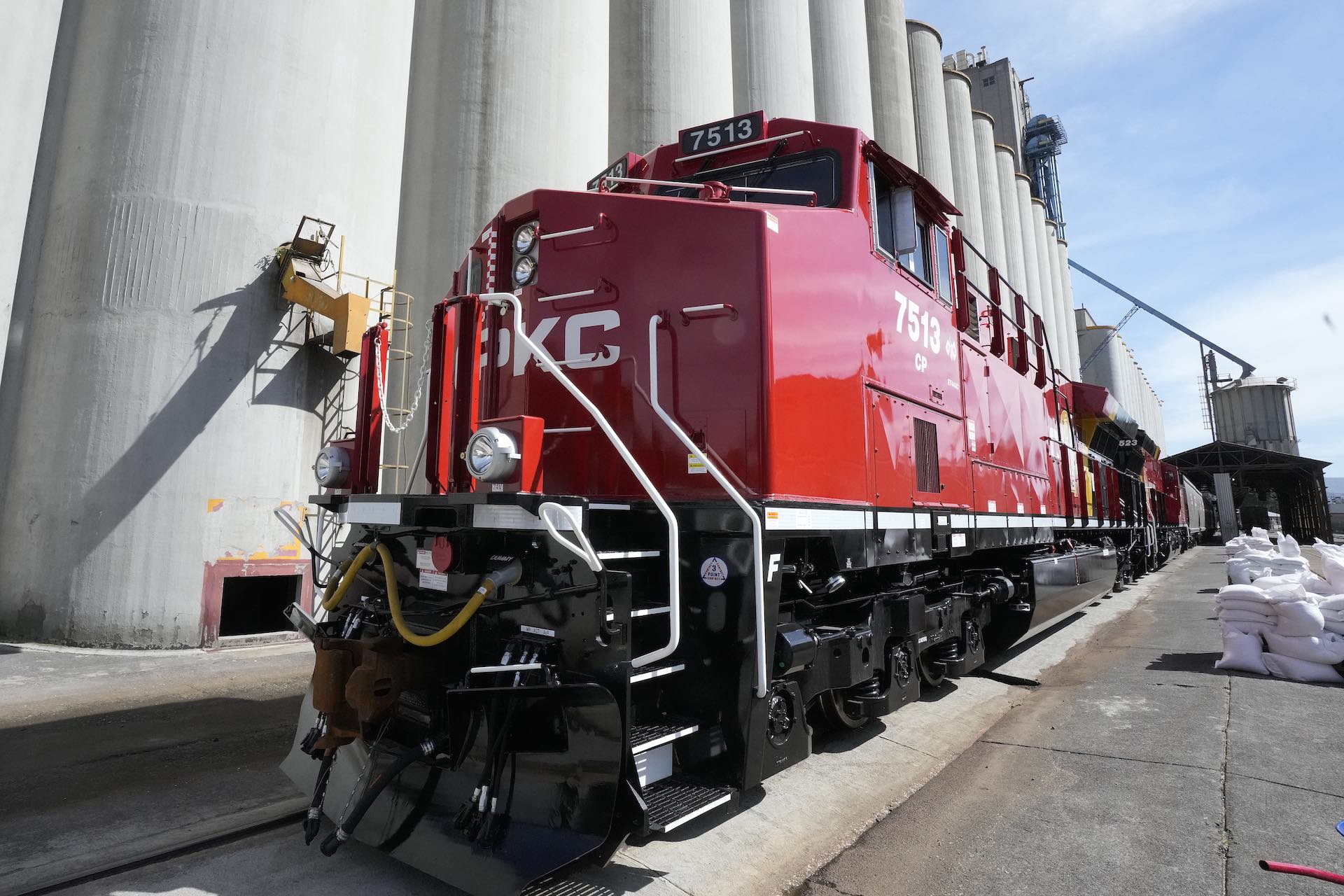 A shot of a shiny, red train locomotive and rail cars next to a row of concrete grain silos.