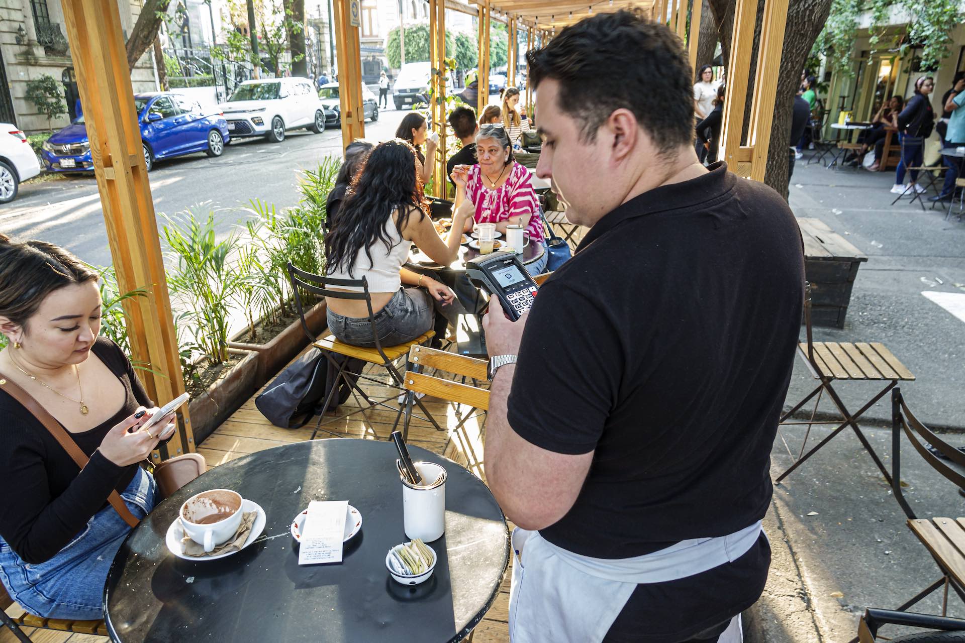 A waiter taking electronic payment from a female customer at an outdoor cafe on a street in Mexico City.