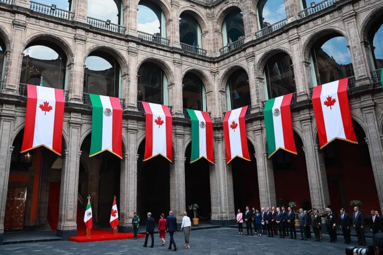 A wide shot of the courtyard at the Palacio Nacional in Mexico City, which is draped with large banners bearing the colours and emblems of the Canadian and Mexican flags. Mark Carney and Claudia Sheinbaum are walking toward a red-carpeted entrance, with other dignitaries standing to their right.