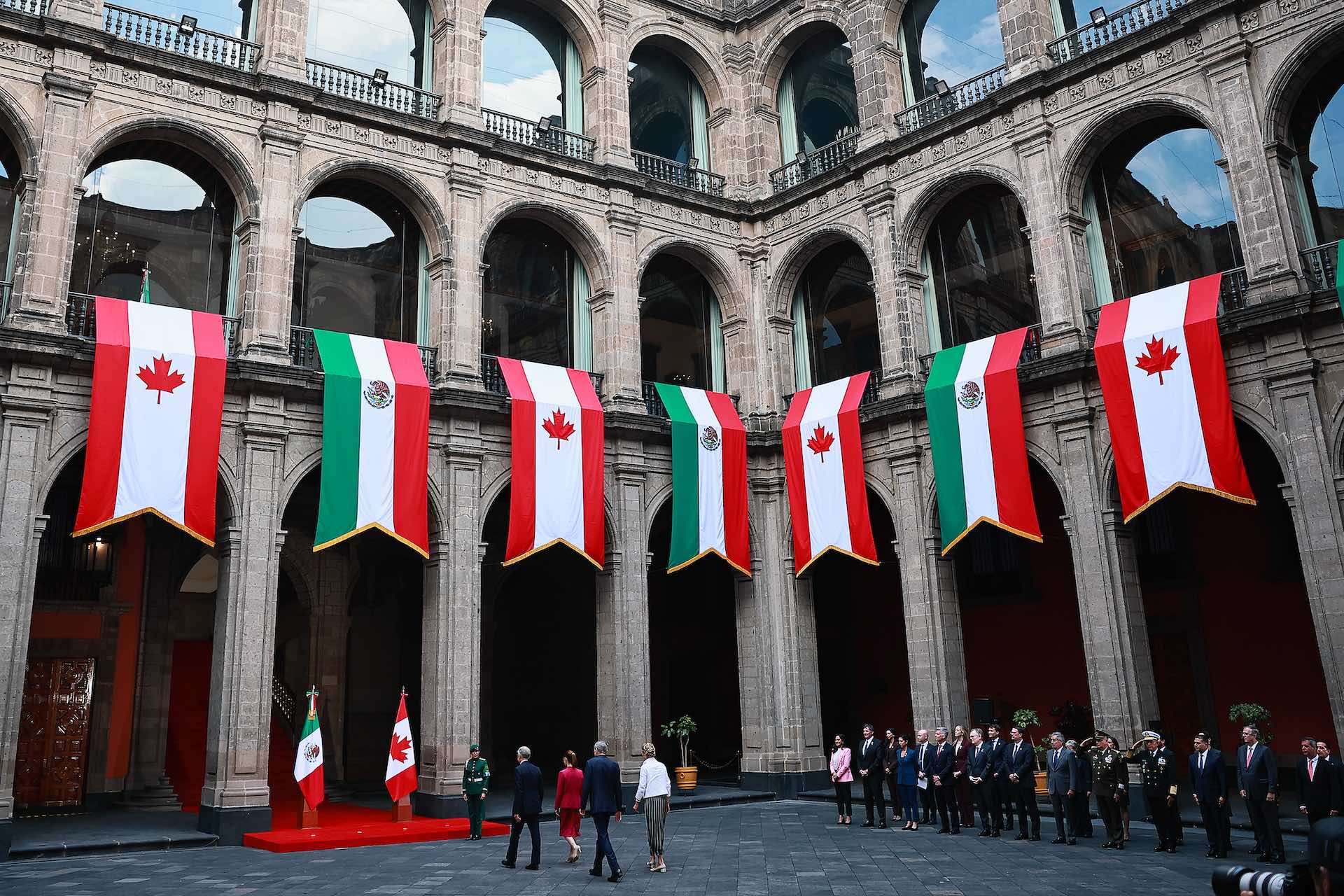 A wide shot of the courtyard at the Palacio Nacional in Mexico City, which is draped with large banners bearing the colours and emblems of the Canadian and Mexican flags. Mark Carney and Claudia Sheinbaum are walking toward a red-carpeted entrance, with other dignitaries standing to their right.
