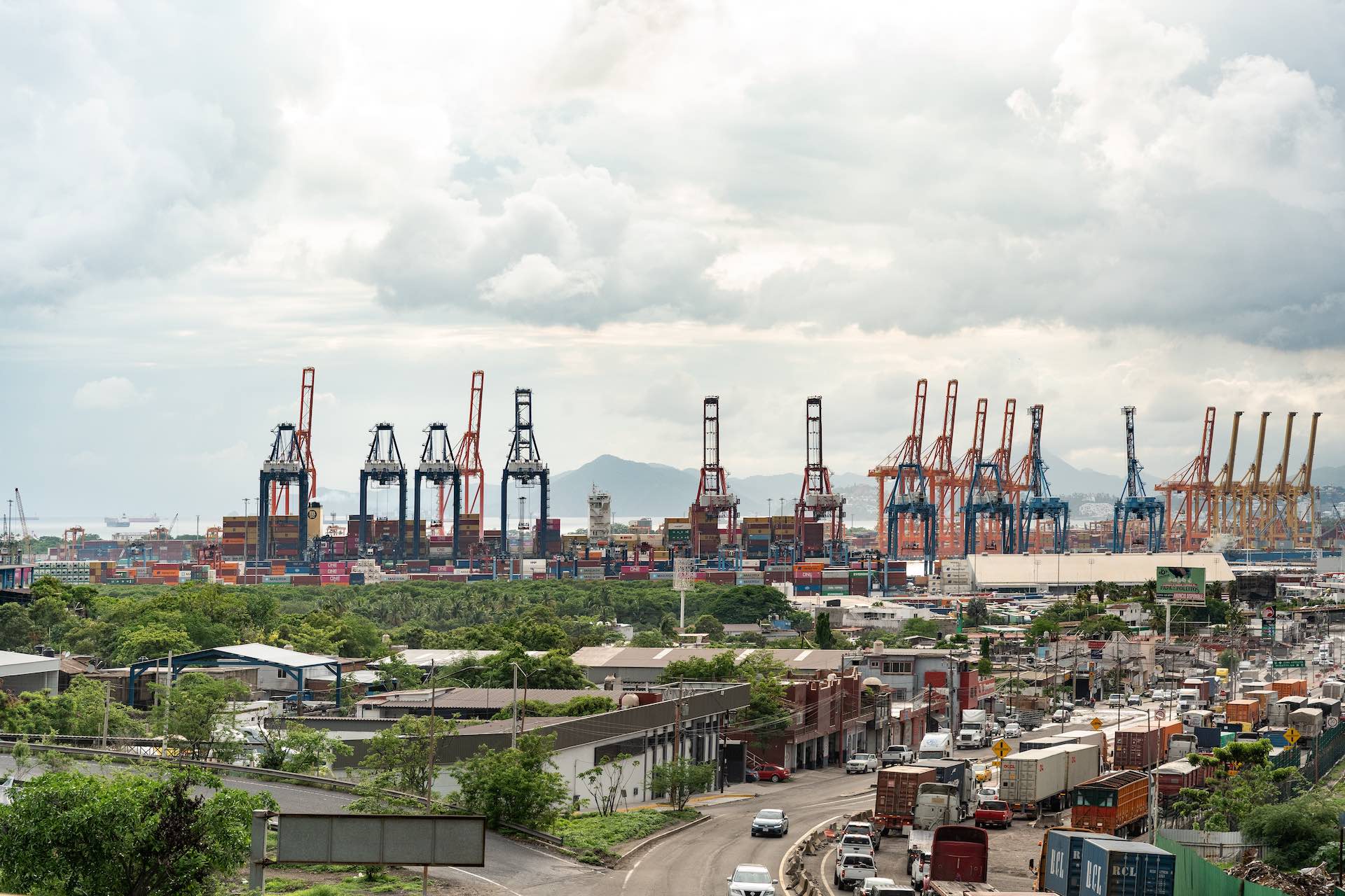 An aerial view of the Port of Manzanillo, Colima, Mexico, showing truck traffic on nearby roads and port facilities.