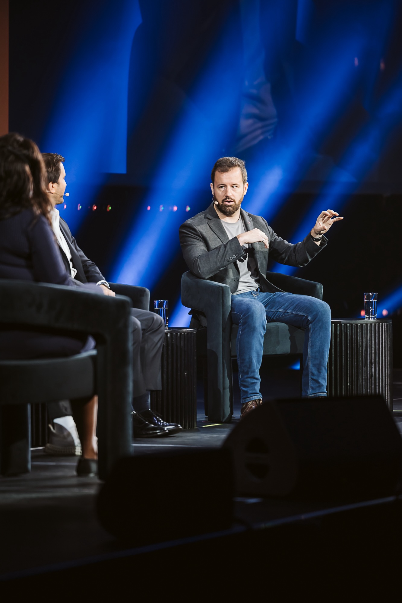 A man sits in a chair on stage and talks to panelists on stage with him.