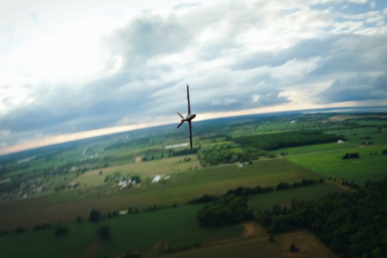 A shot taken from the air of a black, fixed-wing drone flying over farmland. The drone has rolled 90 degrees, so its wings are perpendicular to the horizon.