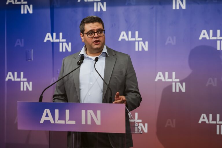 Hypertec CEO Simon Ahdoot in a collared shirt and blazer (no tie) speaks at a podium with the logo for the "All In" conference on it. A backdrop behind him has the logo on it several times over.