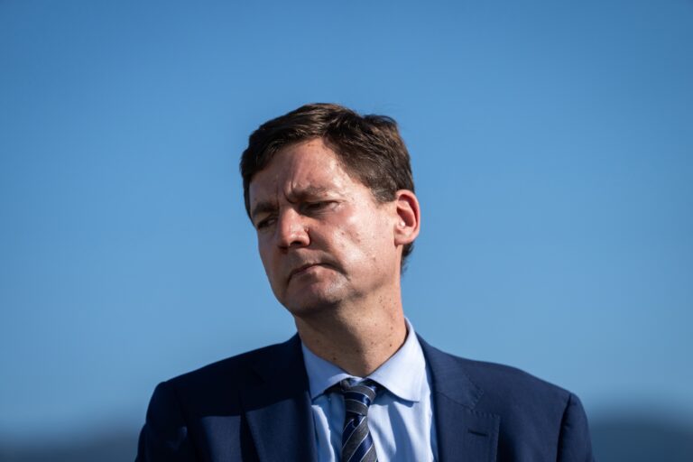 David Eby in a blue suit and striped tie stands against a clear blue sky, looking pensive. The lighting highlights his serious expression, suggesting deep thought.