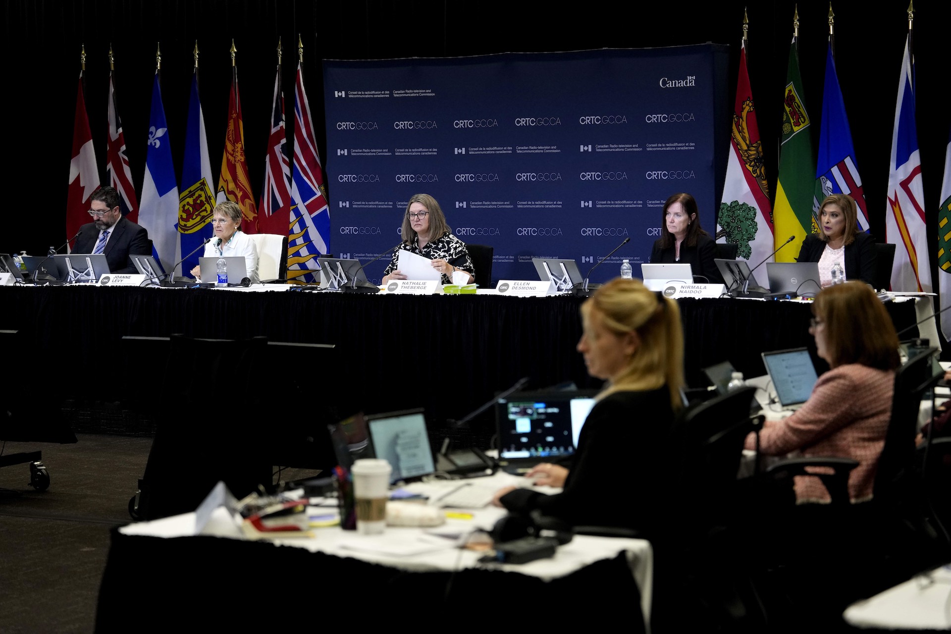 A wide shot of a hearing room with CRTC commissioners seated at the front. There are Canadian and provincial flags behind them.