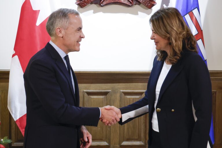Carney and Smith shaking hands in front a wall with wooden wainscoting and the flags of Alberta and Canada.