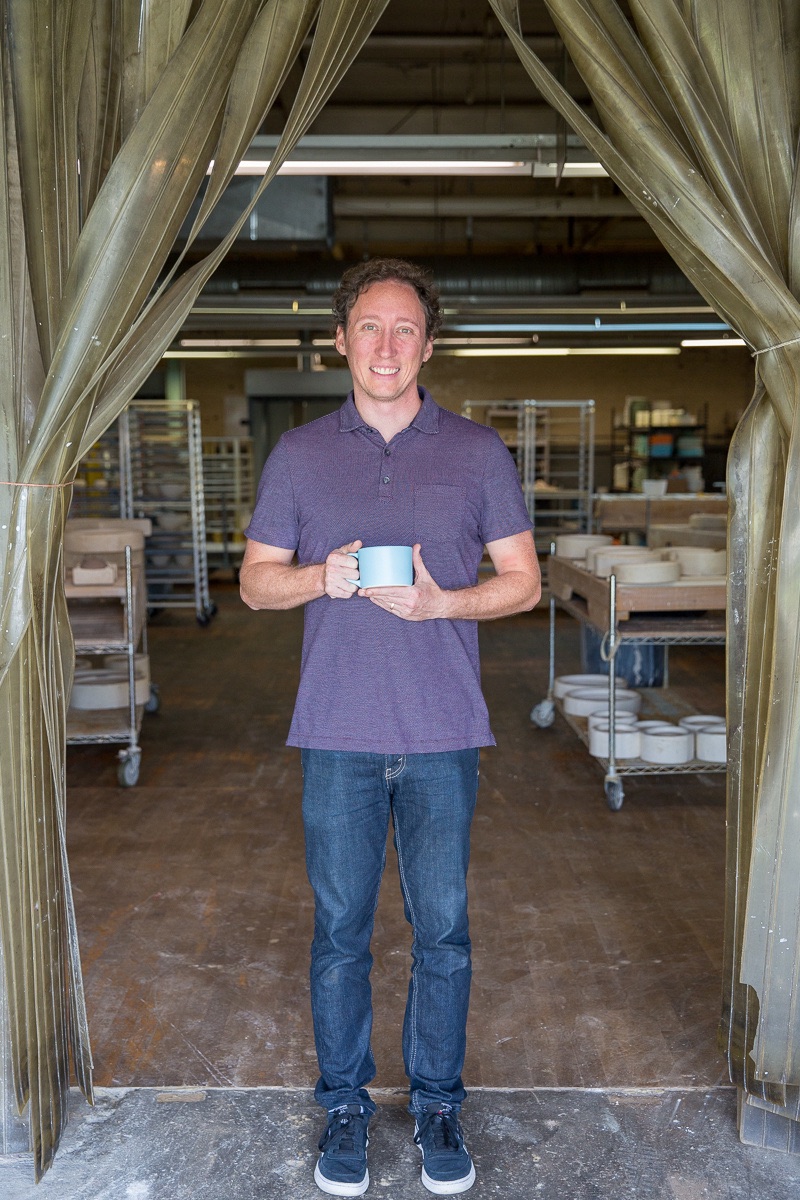 Chris Pence, wearing jeans and a purple golf shirt, in a posed portrait with pottery on carts in the background. He's hold ing a light blue mug in front of him.