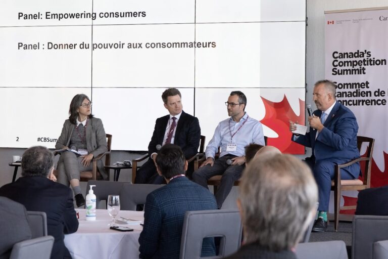 A panel of speakers sits onstage in front of a white screen and a Canadian flag on the left side at the Competition Summit 2025 during the “Empowering Consumers” session, moderated by Vass Bednar, with panelists Geoff White, Sara Eve Levac, Blair Wiley and Maxime Cohen participating in the discussion.