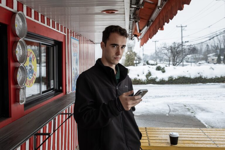 Image of a man standing next to a Slush Puppie stand, holding out his cell phone and looking at the phone. The roadside behind him is covered in snow.