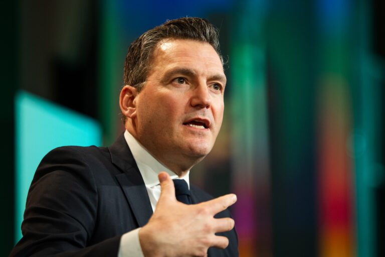 Evan Solomon speaks in front of a blurred multi-coloured background