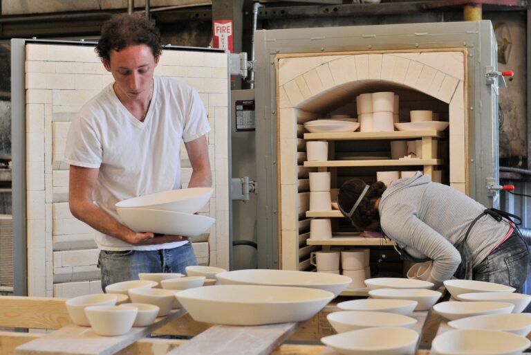 Chris Pence handling serving bowls in the foreground while a female colleague removes dishes from a standup kiln in the background.