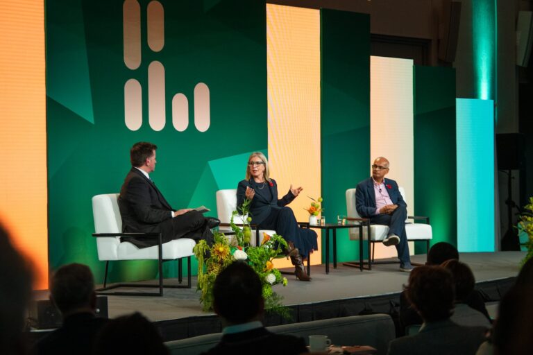 Melanie Woodin sits on stage between a moderator and Vivek Goel, gesturing as she makes a point.