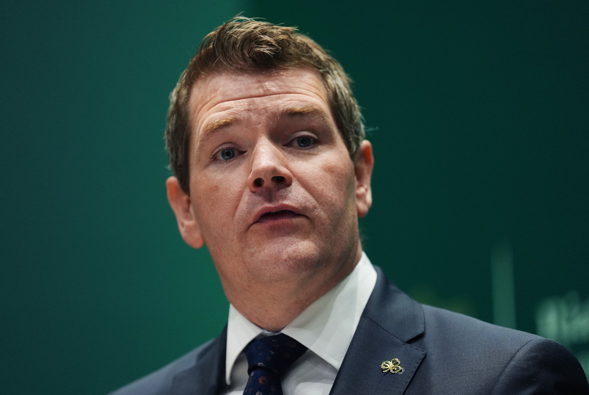 A close-up of Peter Burke against a green backdrop. He's wearing a tie and dark jacket, with a shamrock lapel pin.