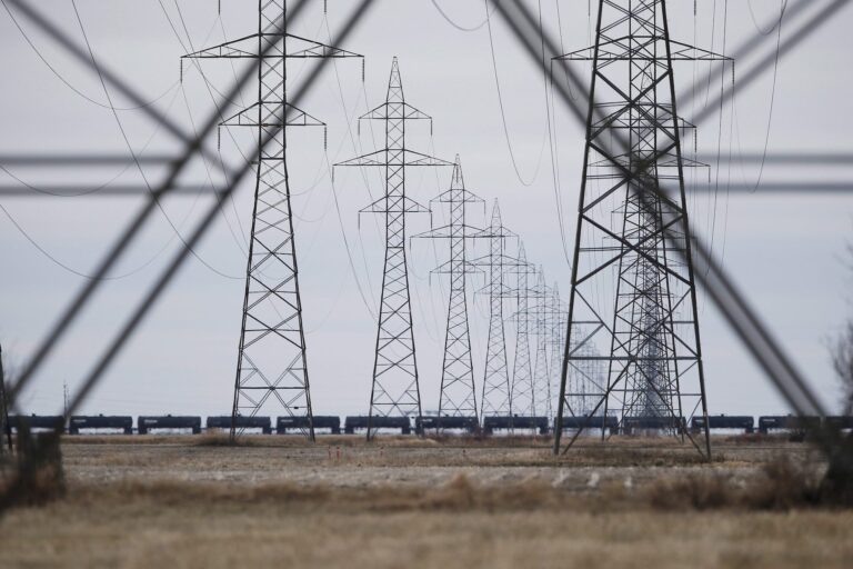 A landscape photo showing high-tension power lines running toward a bleak prairie horizon. There is a row of oil tanker rail cars in the middle of the shot.