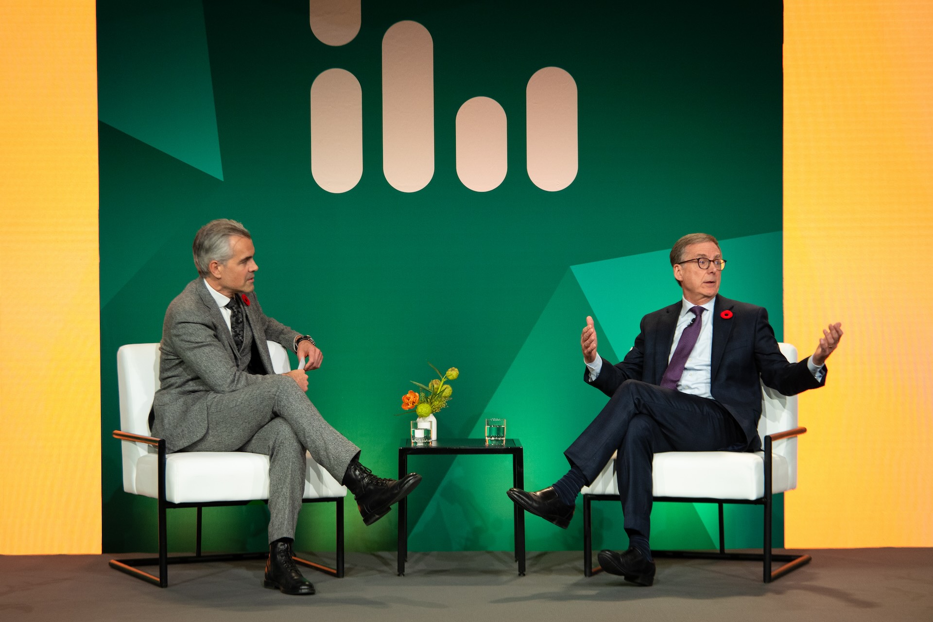 Kevin Carmichael sits onstage, looking at Tiff Macklem, who is gesturing with his arms open as he looks in the opposite direction, speaking to the audience