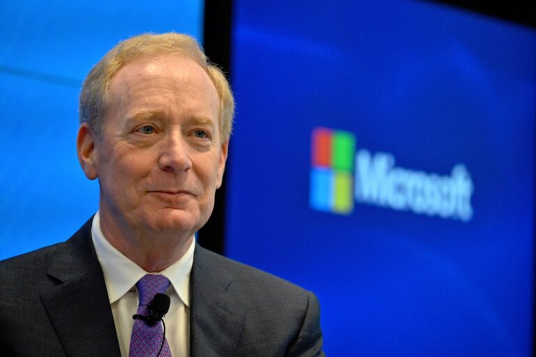A close-up of Brad Smith. He's wearing a suit, and a purple tie with a remote mic attached to it. The Microsoft logo is visible on a large screen behind him.