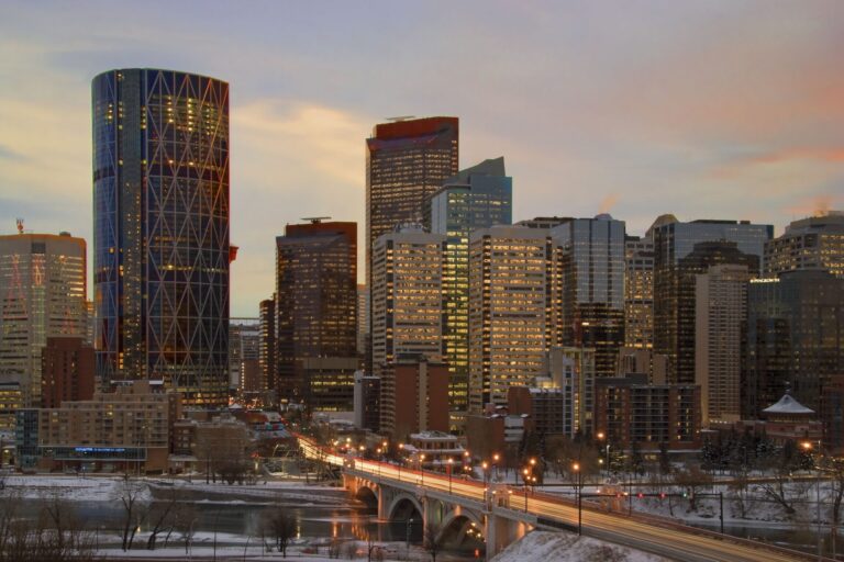 Calgary skyline at dusk with illuminated skyscrapers against a pastel sky. A lit bridge crosses the river, conveying an urban evening atmosphere.
