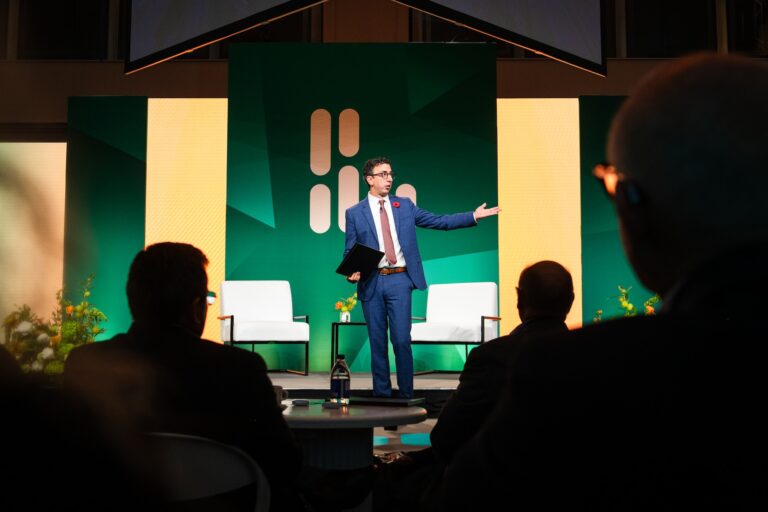 David Skok is in a blue suit, speaking energetically on stage at The Logic Summit, gesturing with one hand. A green-and-yellow geometric backdrop is in the background, while the audience listens intently.