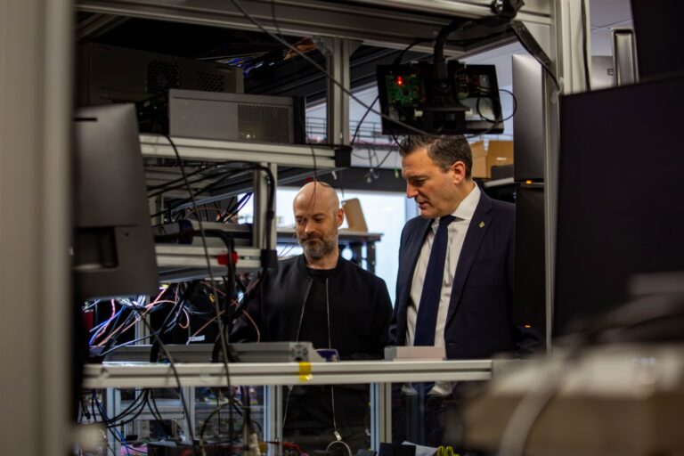 Xanadu CEO Christian Weedbrook and AI Minister Evan Solomon are seen through a mess of electronic equipment and wires on shelving. They are looking down at a part of the shelving and Solomon is saying something.