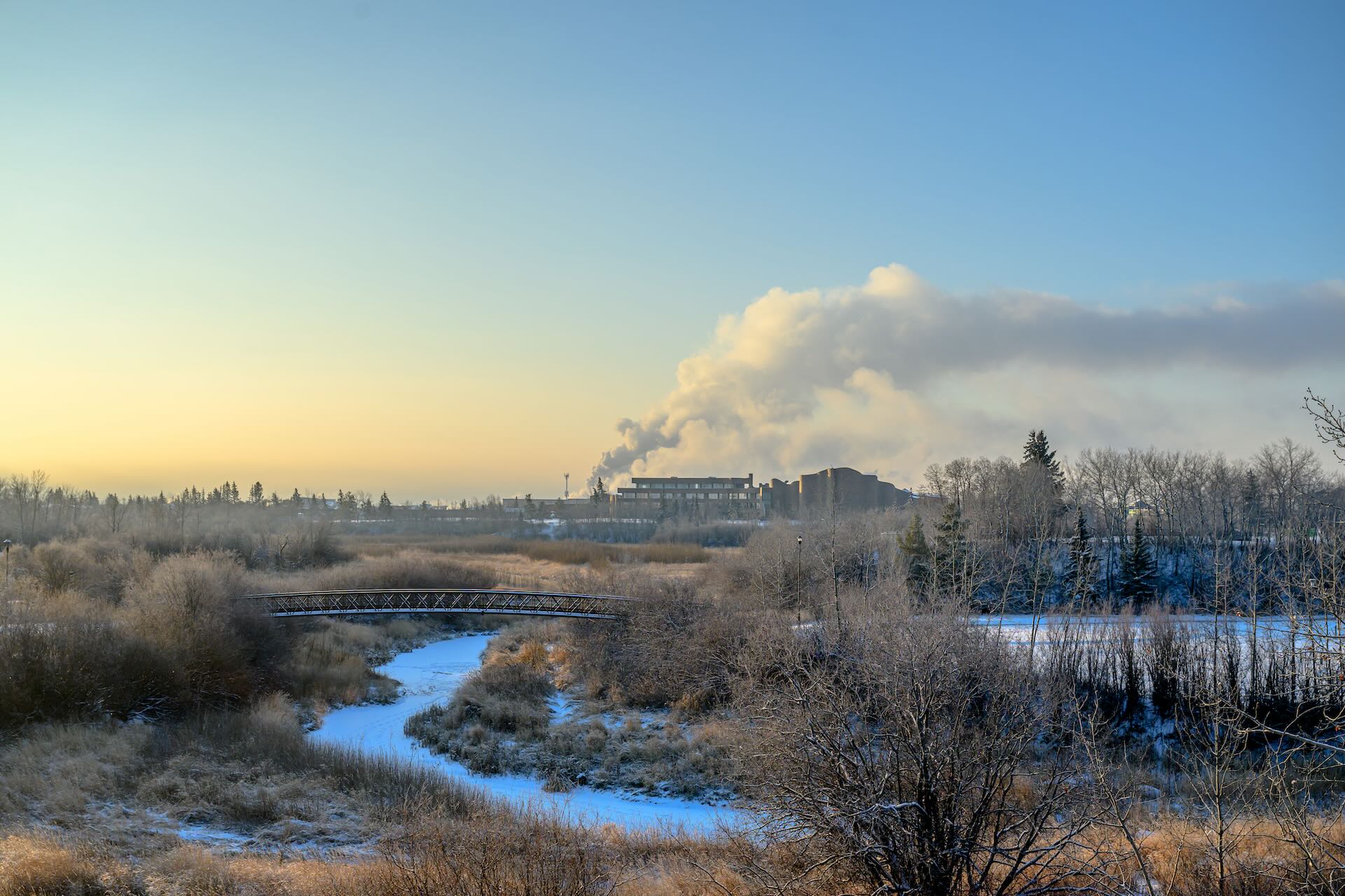 A small bridge crossing over a narrow creek in a field. In the distance are a handful of buildings and a plume of vapour.