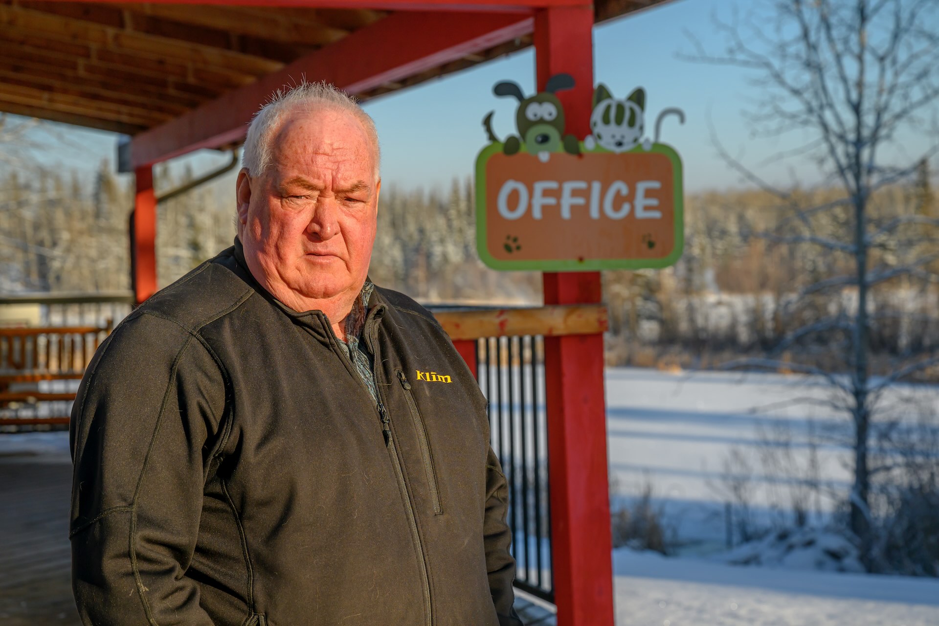 An older man in a black jacket stands in front of a red porch with an "Office" sign decorated with a cat and a dog.
