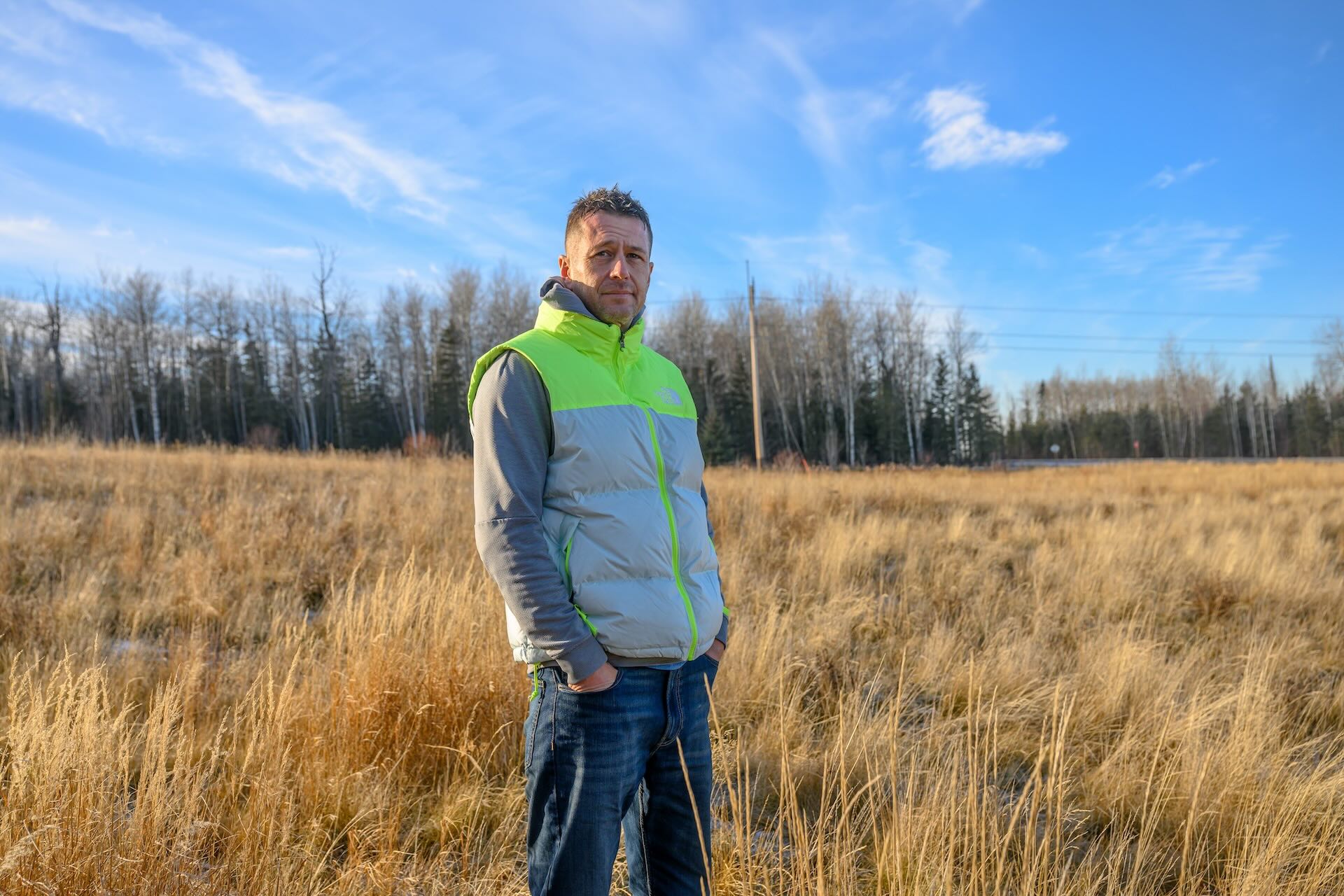 A man in jeans and a hoodie covered by a puffy sleeveless vest with bright yellow highlights stands with his hands in his pockets in a field, with trees and a road in the distance.