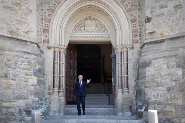 A distant shot of Mark Carney waving his left hand from a flight of stone steps. Behind him are the neo-Gothic arch and tall set of wooden doors of a parliamentary entranceway.