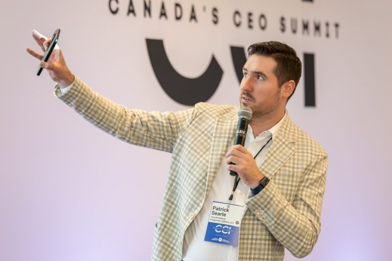 Patrick Searle speaking into a microphone and gesturing with his right hand, which is holding a smartphone. He's wearing a light-toned patterned jacket and an open-collared shirt.