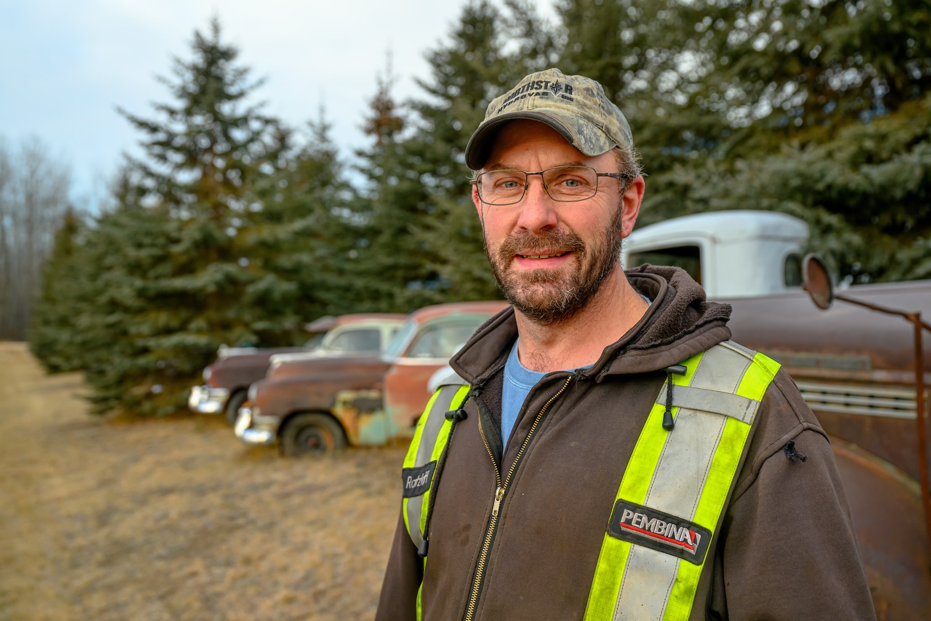 A man in a camo baseball hat and brown zip-up hoodie with a yellow Pembina safety harness on stands in front of a row of rusted cars and trucks in a field